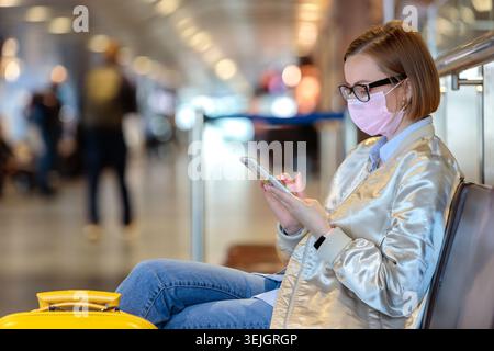 Woman wearing face mask using smartphone inside train station or airport, waiting to board a flight Stock Photo