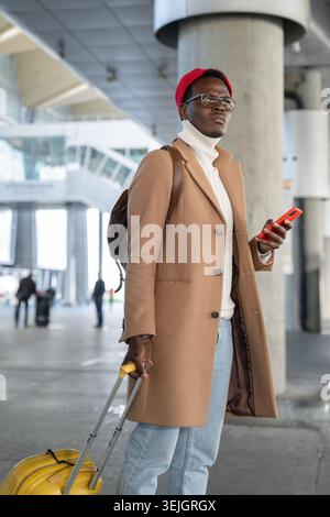 Afro-American traveler man in beige coat, red hat with yellow suitcase holding mobile phone, waiting public transportation in ai Stock Photo