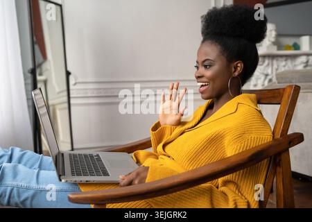 Smiling Afro-American biracial millennial woman in yellow cardigan resting, sitting on armchair, talking in video chat with her Stock Photo