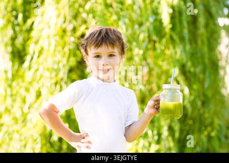Cute child boy drinks healthy green smoothie with straw in a jar mug against the background of greenery outdoor. summer detox dr Stock Photo