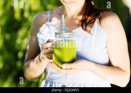 Woman hand holding healthy green smoothie with straw in a jar mug against the background of greenery. summer detox drinks. Jam G Stock Photo