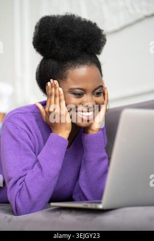 Smiling Afro-American millennial woman in purple sweater lying on couch, looking at camera webcam and talking on a video call or Stock Photo
