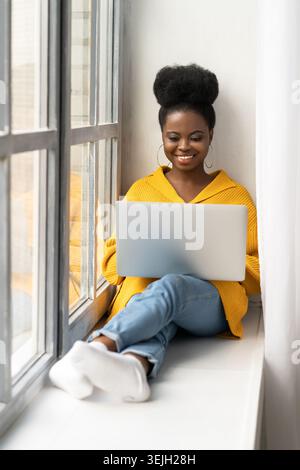 Smiling Afro-American biracial woman in yellow cardigan sitting on windowsill, distance working on laptop, chatting and talking Stock Photo