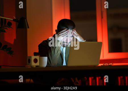 Pensive young businessman at night office work at laptop computer exhausted and tired of deadlines and overwork. Frustrated programmer trying to finis Stock Photo