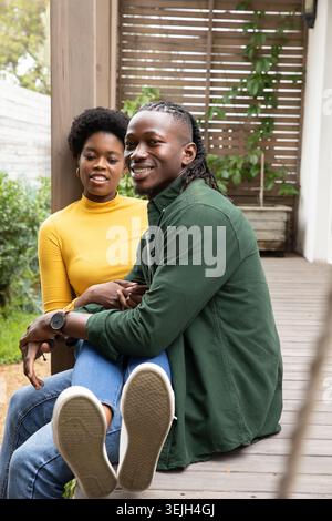 African American couple sitting on deck hugging wearing yellow top and sneakers near planter vines. Romantic, affection, cozy, outdoor, rustic, leisur Stock Photo
