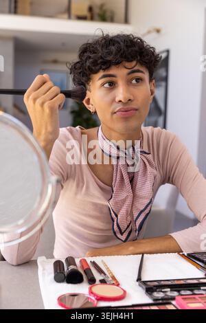 Man applying facial makeup at apartment table using brush beside round mirror and eyeshadow palette. Cosmetics, grooming, vanity Stock Photo