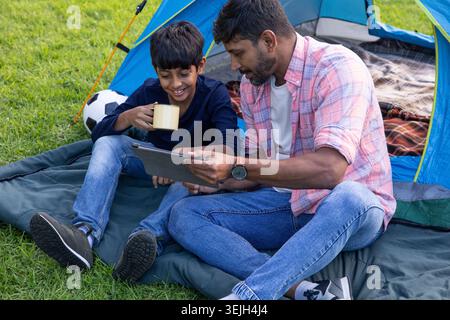 Indian father and son sitting near blue tent beside soccer ball, using tablet, holding yellow mug. Adventure, bonding, leisure, nature, outdoor, playf Stock Photo
