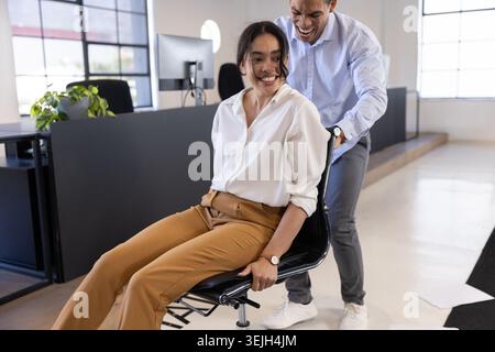 Diverse coworkers in office pushing rolling chair near monitors, desk partitions and potted plant. Collaboration, teamwork, prod Stock Photo