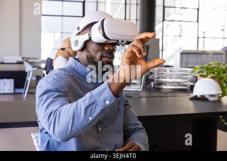 Diverse coworkers wearing VR headset interacting virtually at modern office by laptop and hard hat. Technology, collaboration, i Stock Photo
