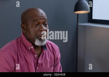 Senior African American man sitting against grey wall in lounge area under black pendant lamp. Portrait, mature, contemporary, minimalist, relaxation, Stock Photo