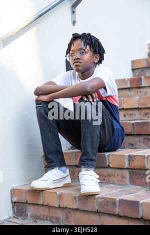 African American boy sitting on brick steps under metal handrail wearing eyeglasses and T shirt. Urban, childhood, contemplation Stock Photo