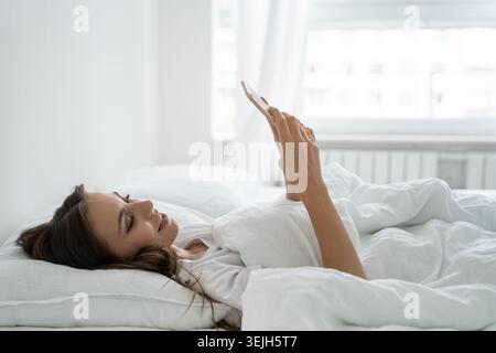 Smiling young woman using mobile smart phone, lying and relaxing in white comfortable bed at home Stock Photo