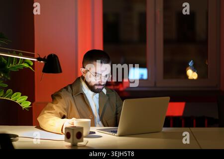 Serious young man looking at laptop computer drink coffee at workplace staying in office at dark late night. Male designer or web developer overworkin Stock Photo