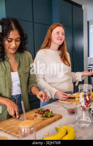 Diverse female friends chopping strawberries and slicing bananas by blender on kitchen countertop. Cooking, collaboration, welln Stock Photo