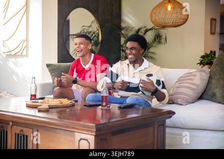 Diverse male friends laughing while holding soccer ball and eating pizza on living room sofa. Casual, friendship, relaxation, mo Stock Photo