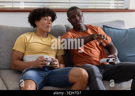 Diverse male friends sitting on living room sofa by blinds holding white gamepads with blue cushion. Casual, entertainment, leis Stock Photo