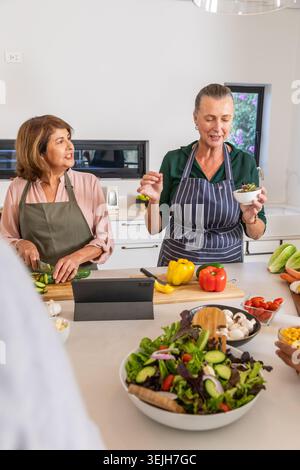 Senior female friends wearing aprons chopping cucumber and tossing greens in kitchen using tablet. Cooking, collaboration, conte Stock Photo