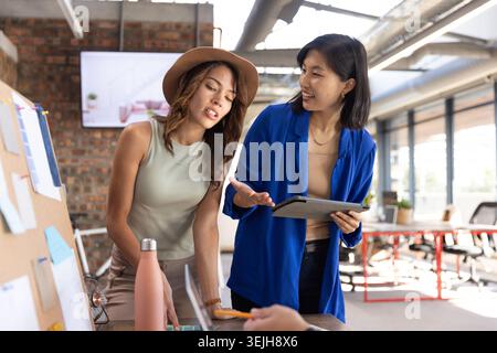 Diverse female colleagues wearing blazer collaborating over cork board at office desk with tablet. Collaboration, teamwork, inno Stock Photo