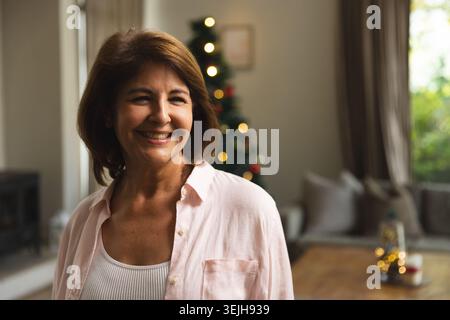 Woman smiling while standing next to sofa in living room with Christmas tree lights, copy space. Festive, cozy, holiday, interio Stock Photo