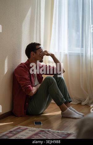 Upset depressed young man sitting on floor at home, received unexpected bad news over phone Stock Photo