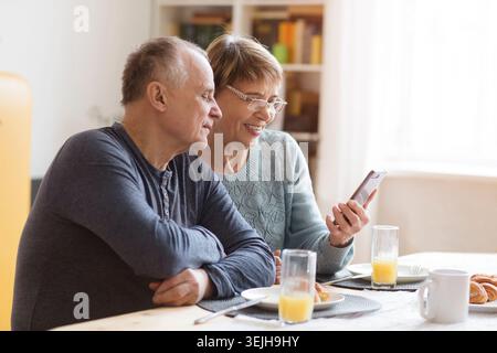 Senior couple eating breakfast and using smartphone at home Stock Photo
