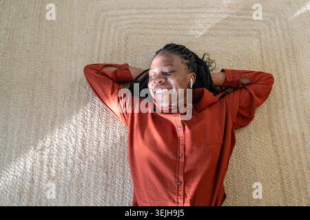 Carefree happy African American woman lying on floor relaxing listening to favorite music in pods. Stock Photo