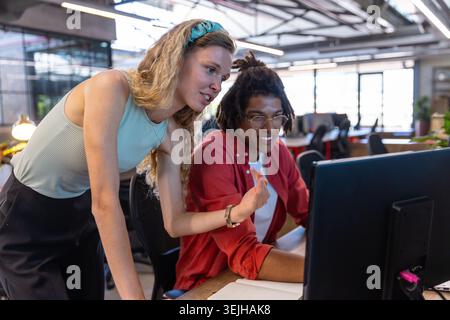 Diverse coworkers collaborating at desk in open plan office using monitor, keyboard, notebook, pen. Collaboration, teamwork, pro Stock Photo