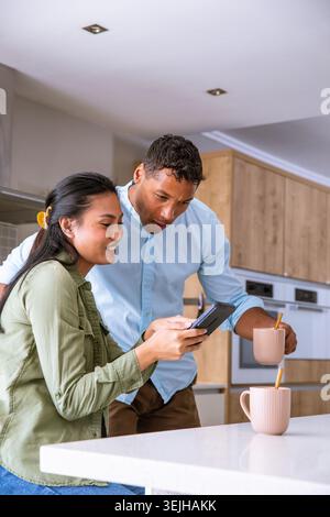 Diverse couple standing by island countertop in modern kitchen holding smartphone and pink mugs. Contemporary, lifestyle, minimalism, domestic, couple Stock Photo