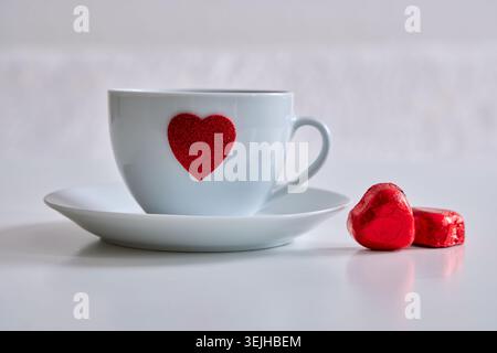 A white coffee cup with a red heart on a saucer next to two heart-shaped chocolates. Stock Photo