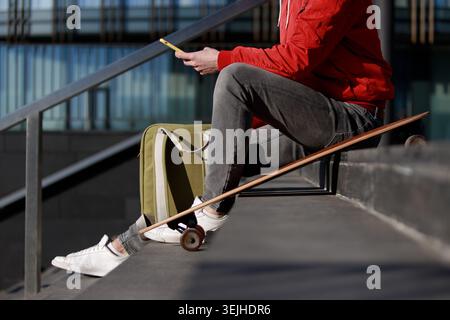 Stylish man longboarder in casual clothes using smartphone, resting on the steps, sitting with longboard, skateboard outdoors. Urban, subculture, skat Stock Photo