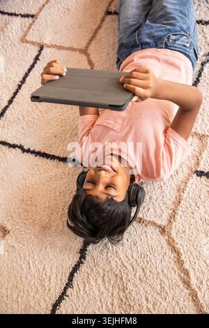 Asian child lying on beige rug at home holding tablet wearing pink shirt and black headphones. Playful, relaxation, leisure, mod Stock Photo