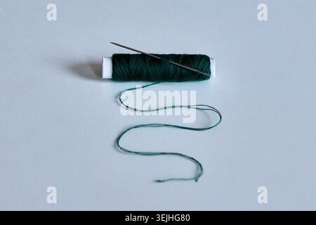 A single spool of dark green sewing thread with a metal needle and loose wavy fiber on a white background. Stock Photo