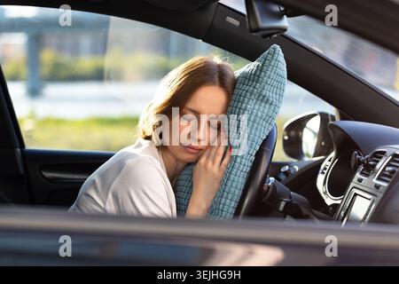 Tired young woman driver asleep on pillow on steering wheel, resting after long hours driving a car. Fatigue. Sleep deprivation. Stock Photo