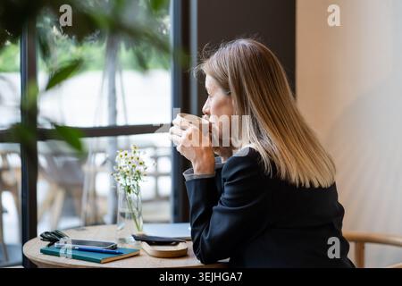 Pensive positive businesswoman drinking coffee and thinking about new work plans in cozy cafe. Stock Photo