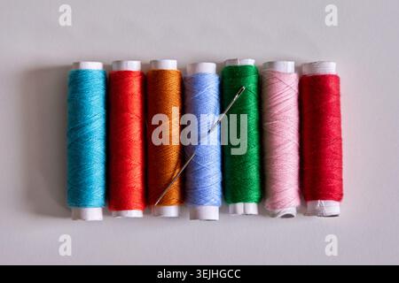 A row of colorful sewing thread spools with a metal needle on a plain white background. Stock Photo