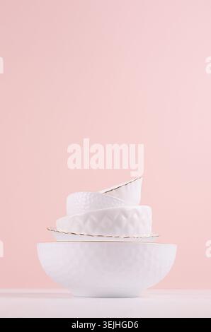 White elegant plates in stack with thin golden bezel, bizarre texture on white wooden table, pastel pink wall, vertical. Modern Stock Photo
