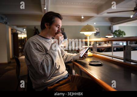 Handsome pensive man enjoying aromatic coffee and scrolling smartphone while relaxing in cafe space of library with co-working area. Thoughtful male c Stock Photo
