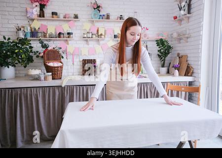 Woman preparing festive Easter table setting with flowers, cakes and painted eggs, laying table with a white tablecloth Stock Photo