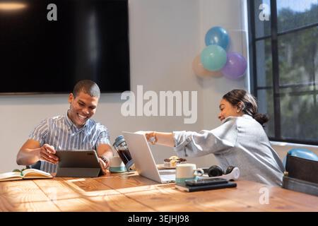 Diverse male coworkers collaborating on laptop at conference table with muffins and pastel balloons. Collaboration, teamwork, mo Stock Photo