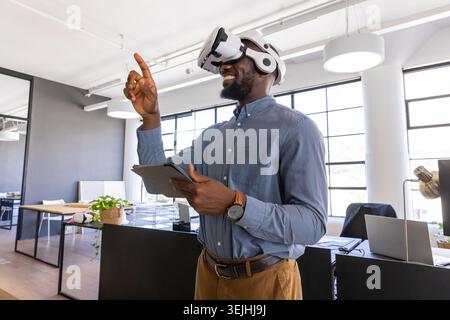 African American man standing in office wearing VR headset and interacting with tablet near laptops. Futuristic, technology, innovation, workspace, di Stock Photo