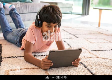 Indian boy lying on beige and black geometric rug in living room using tablet with headphones. Tech, leisure, relaxation, modern Stock Photo