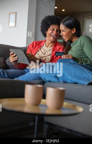 Diverse female friends leaning together on gray couch at home, browsing tablet with credit card. Collaboration, friendship, leis Stock Photo