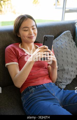 Asian woman reclining on dark gray sofa in living room holding black smartphone and smiling. Relaxation, leisure, comfort, techn Stock Photo