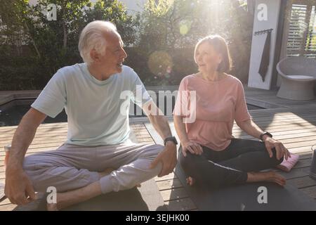 Senior couple practicing seated yoga on yoga mats on wooden deck next to pool with smartwatches. Wellness, serenity, leisure, ou Stock Photo