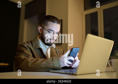 Focused young front end developer or qa engineer testing app on smartphone workloaded, work overtime in empty coworking space before project deadline. Stock Photo