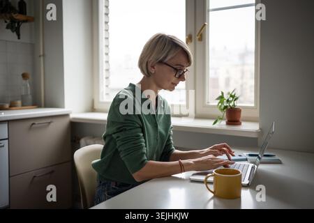 Relaxed middle aged business woman comfortably working typing on laptop online in kitchen. Stock Photo