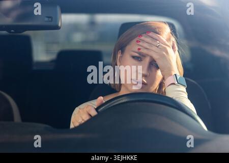 Exhausted young woman driver sitting in her car, feeling emotional burnout after work, looking at camera, touching her forehead. Stock Photo