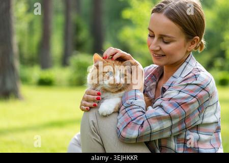 Close up of smiling woman in checked shirt hugging and embracing with tenderness and love domestic ginger cat, stroking on the head, outdoors in sunny Stock Photo