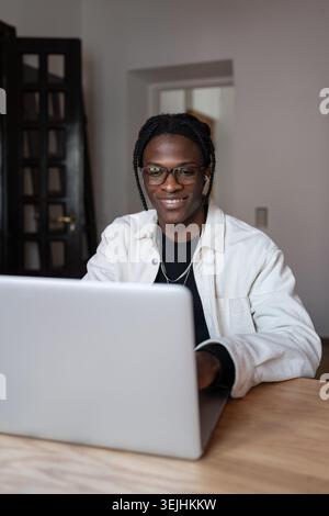 Cheerful charismatic African American man freelancer typing on laptop keyboard sits at wooden table. Smiling satisfied guy using computer for remote w Stock Photo