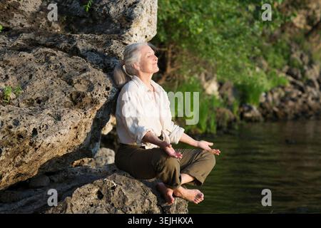 Summer lifestyle portrait senior woman with gray hair relaxes sitting on rocks on the seashore. Enjoying the little things. spen Stock Photo